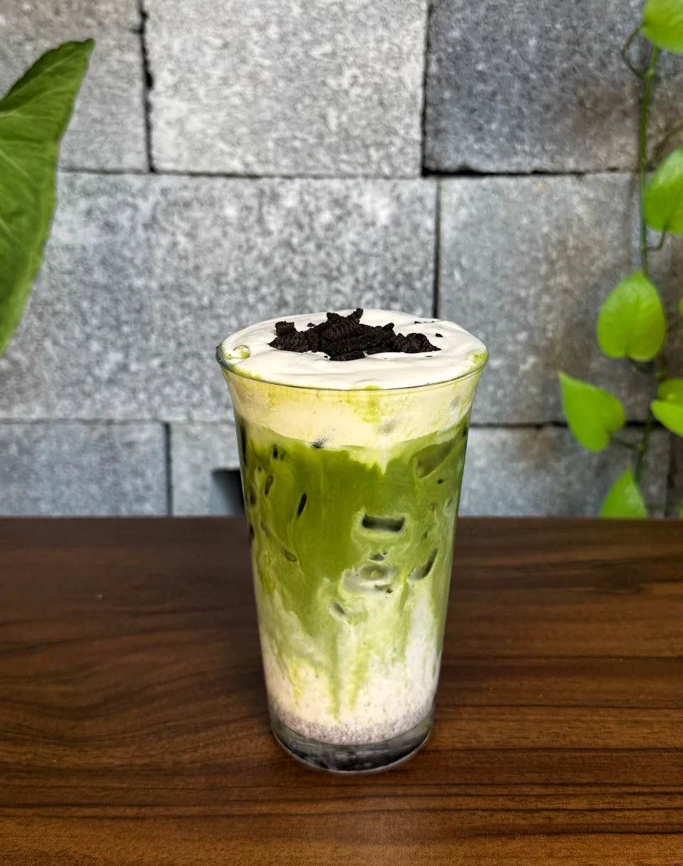 Ly matcha latte với lớp kem cheese trắng và Oreo nghiền rắc bên trên, đặt trên bàn gỗ óc chó.